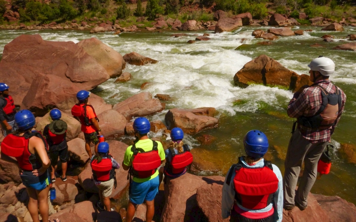 A group of people wearing safety gear stand on the shore of a river, looking out over the water.